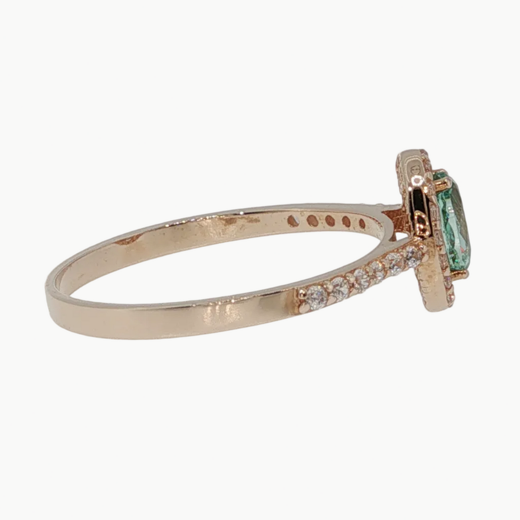 A rose gold ring with a small emerald-cut green gemstone and a row of clear accent stones on the band, shown on a light gray reflective surface.
